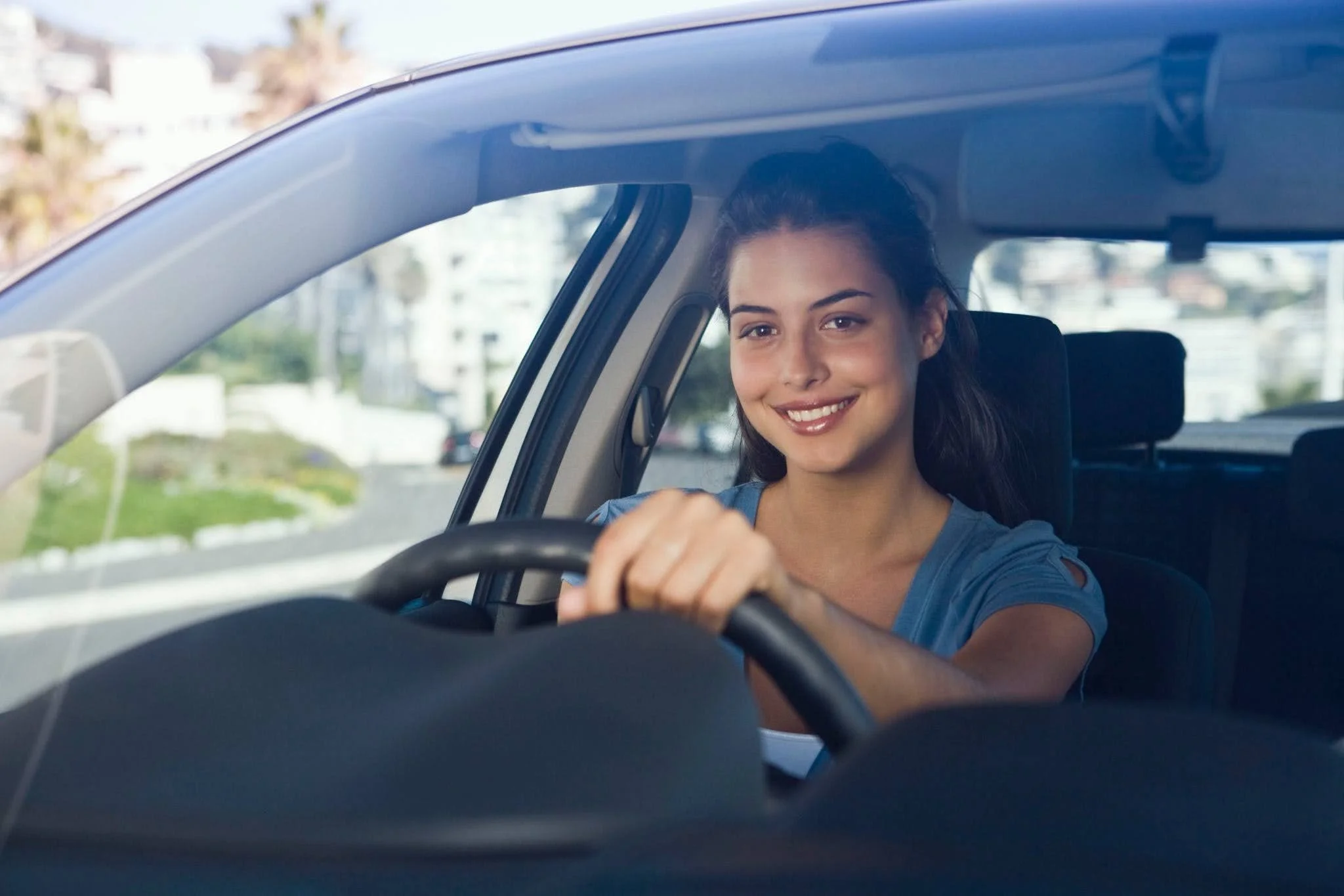 A woman with long dark hair and a blue shirt smiling while sitting behind the wheel of a car during daytime.