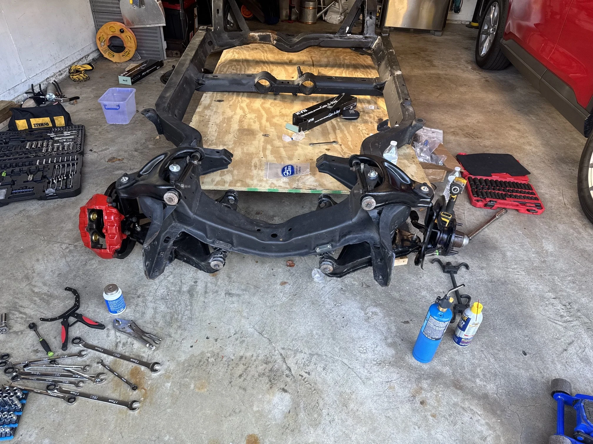 A garage workspace with a partially disassembled car frame on a plywood surface, surrounded by tools, spray bottles, a red brake caliper, a water bottle, and various mechanical parts and equipment.