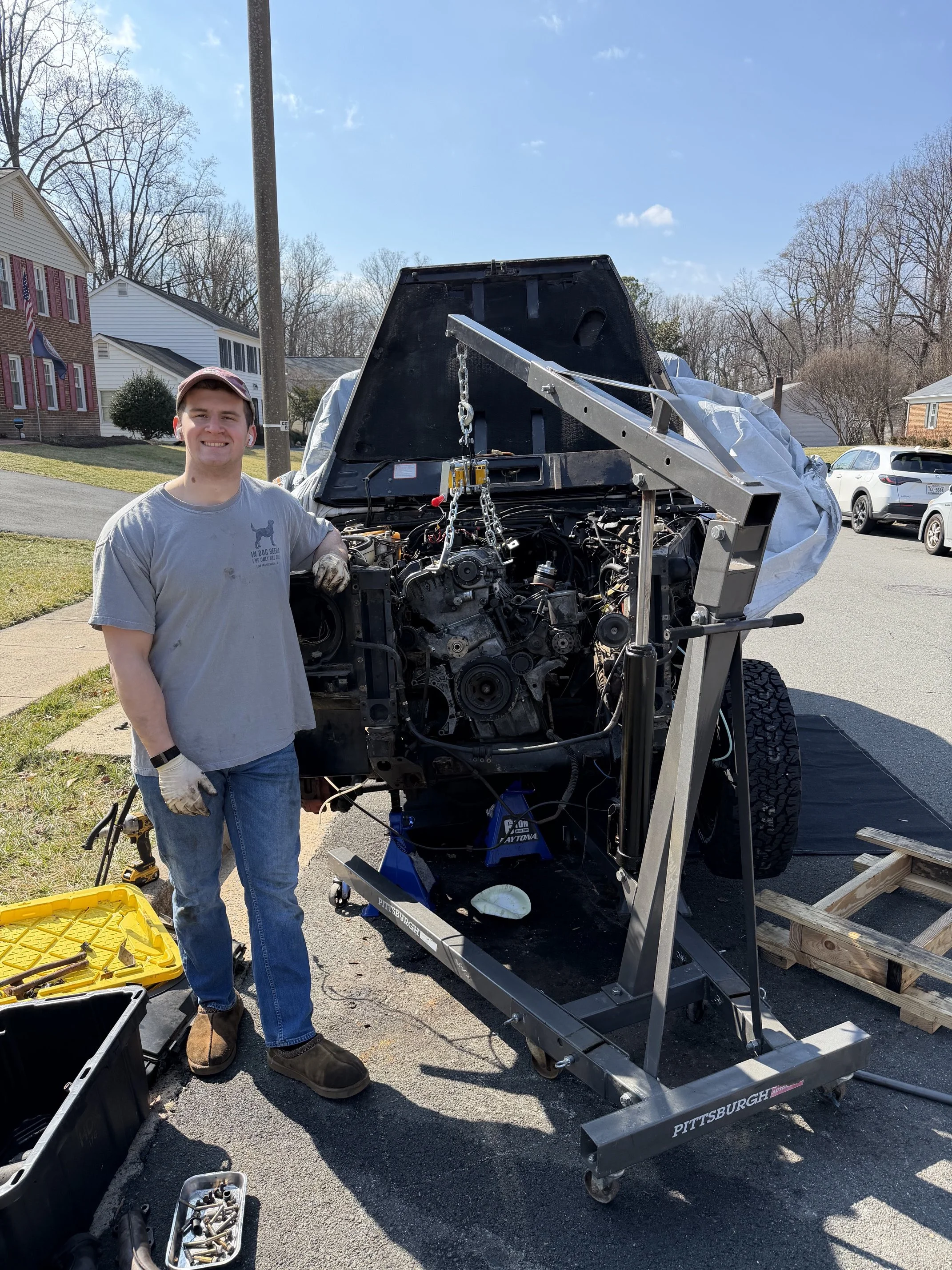 Young man smiling in work clothes, standing beside a partially disassembled vehicle on a wheeled frame outside on a sunny day, with houses and trees in the background.