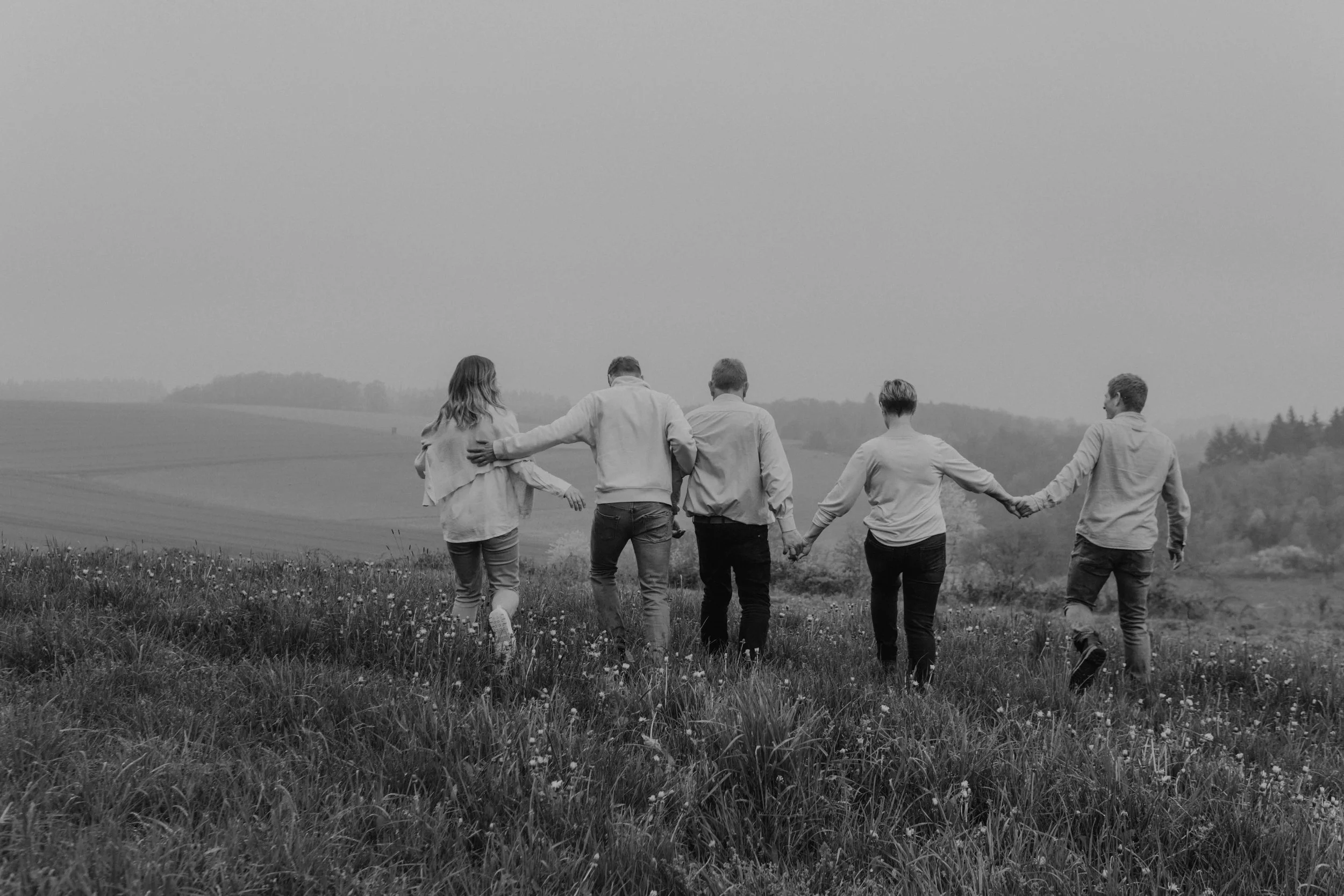 Authentische Familienfotografie in Nordhessen – Eine Gruppe von sechs Menschen, die sich an den Händen halten, läuft durch eine ländliche Landschaft mit Feldern und Bäumen – Familienmomente.