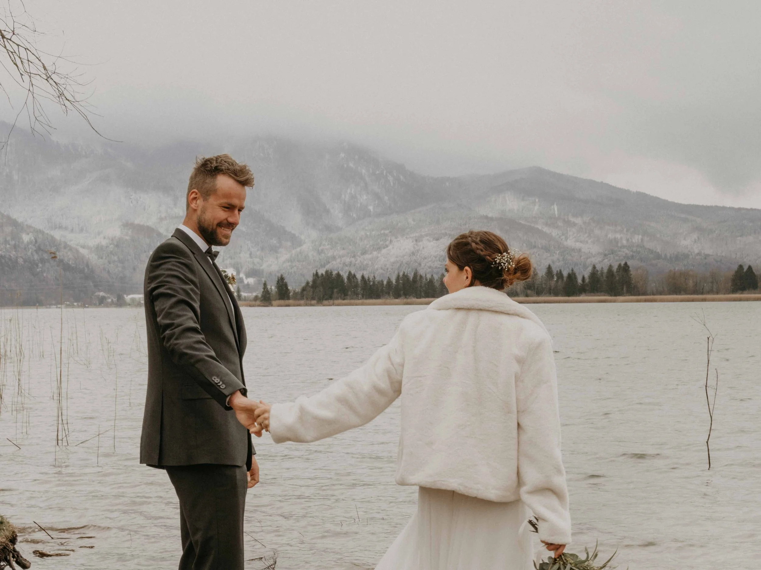 Editorial Hochzeit in Bayern, bad Tölz am Eibsee – Ein Brautpaar hält am Ufer eines Sees Händchen, im Hintergrund Berge und bewölkter Himmel.