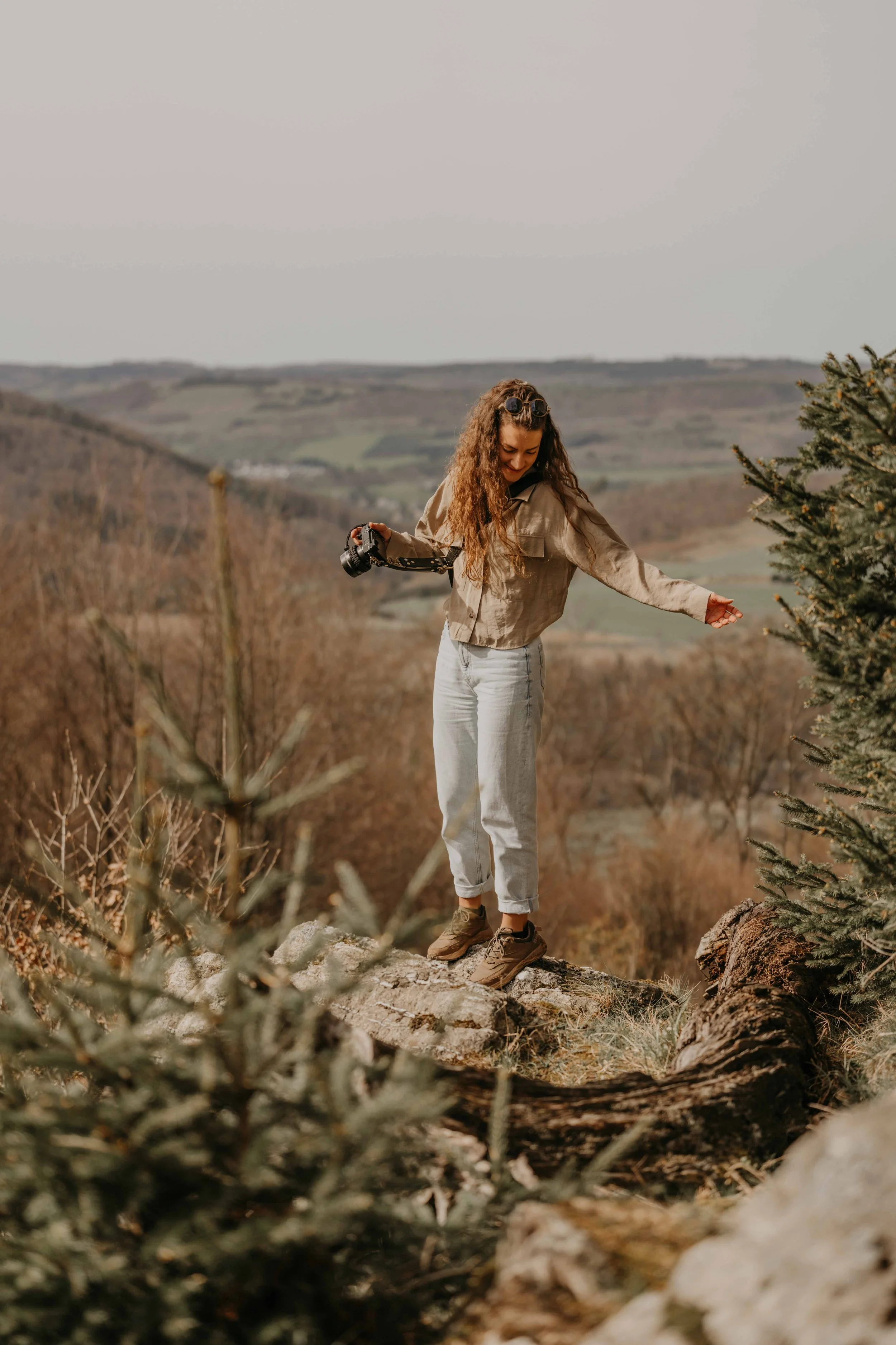 Jara-Eileen Lenz, Hochzeitfotografin Deutschland und Weltweit –  Junge Frau mit Kamera steht auf einem Baumstamm in einer Naturlandschaft im Sauerland an den Bruchhäuser Steinen, lachend, ländliche Umgebung.