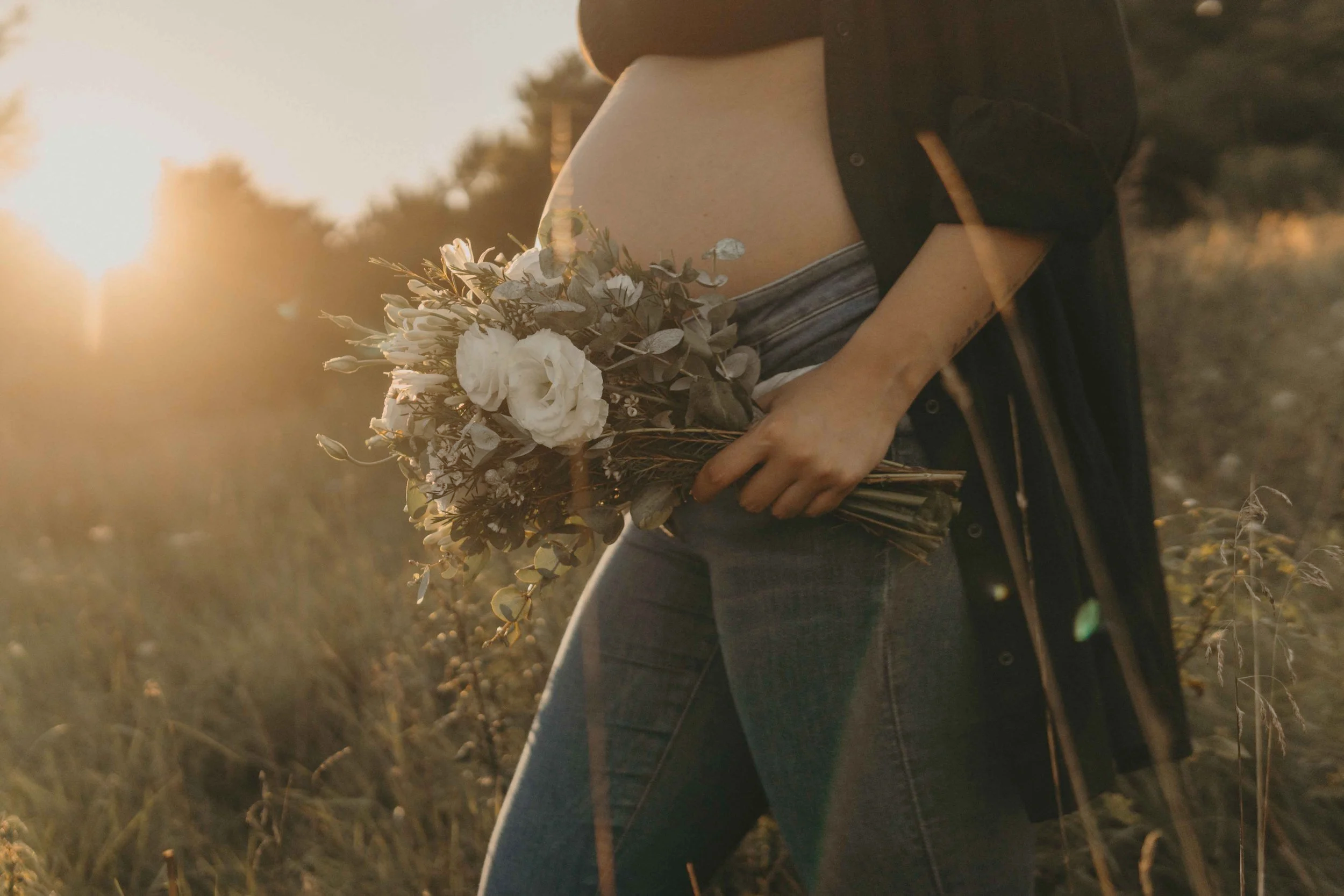 Babybauchshooting Hamburg – werdende Mutter hält zärtlich ihren Bauch und Blumen im Sonnenuntergang.