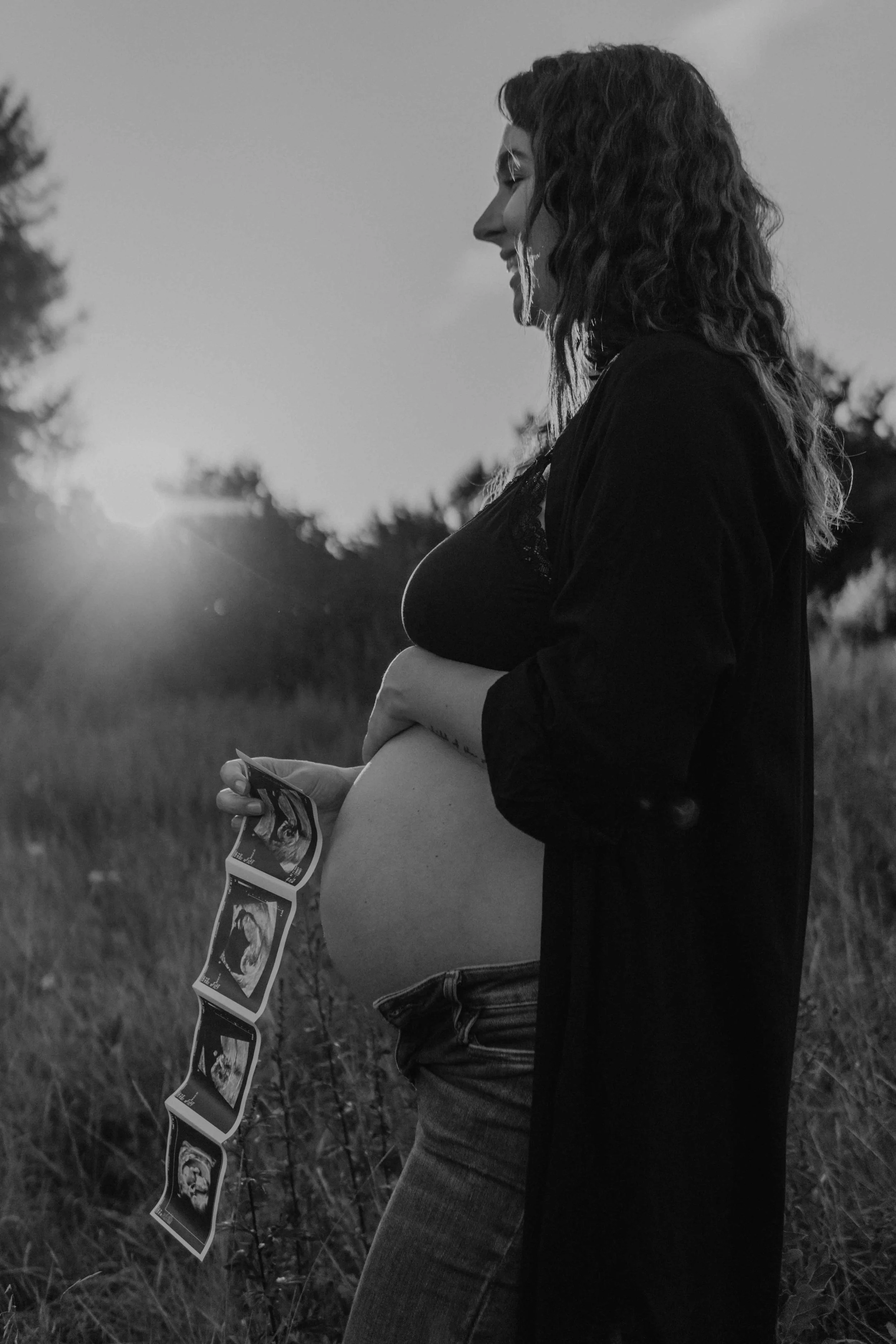 Babybauchshooting Hamburg – werdende Mutter hält zärtlich ihren Bauch und das Ultraschallbild im Sonnenuntergang.