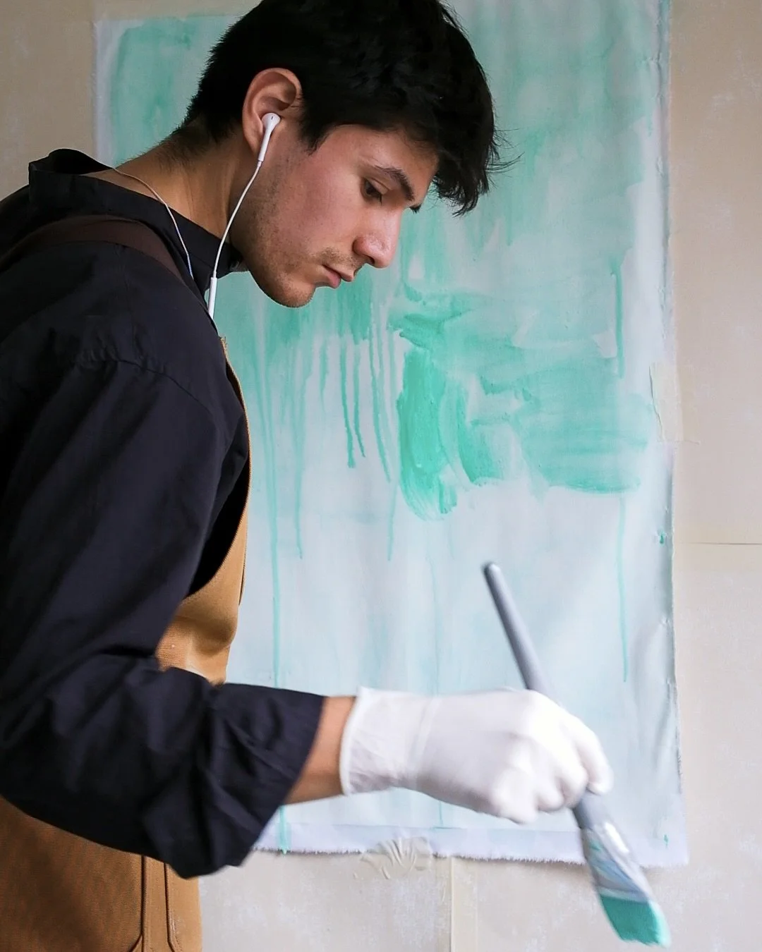 Un jeune homme en train de peindre avec un pinceau vert devant un tableau ou un mur avec de la peinture verte, portant des gants blancs et des écouteurs.