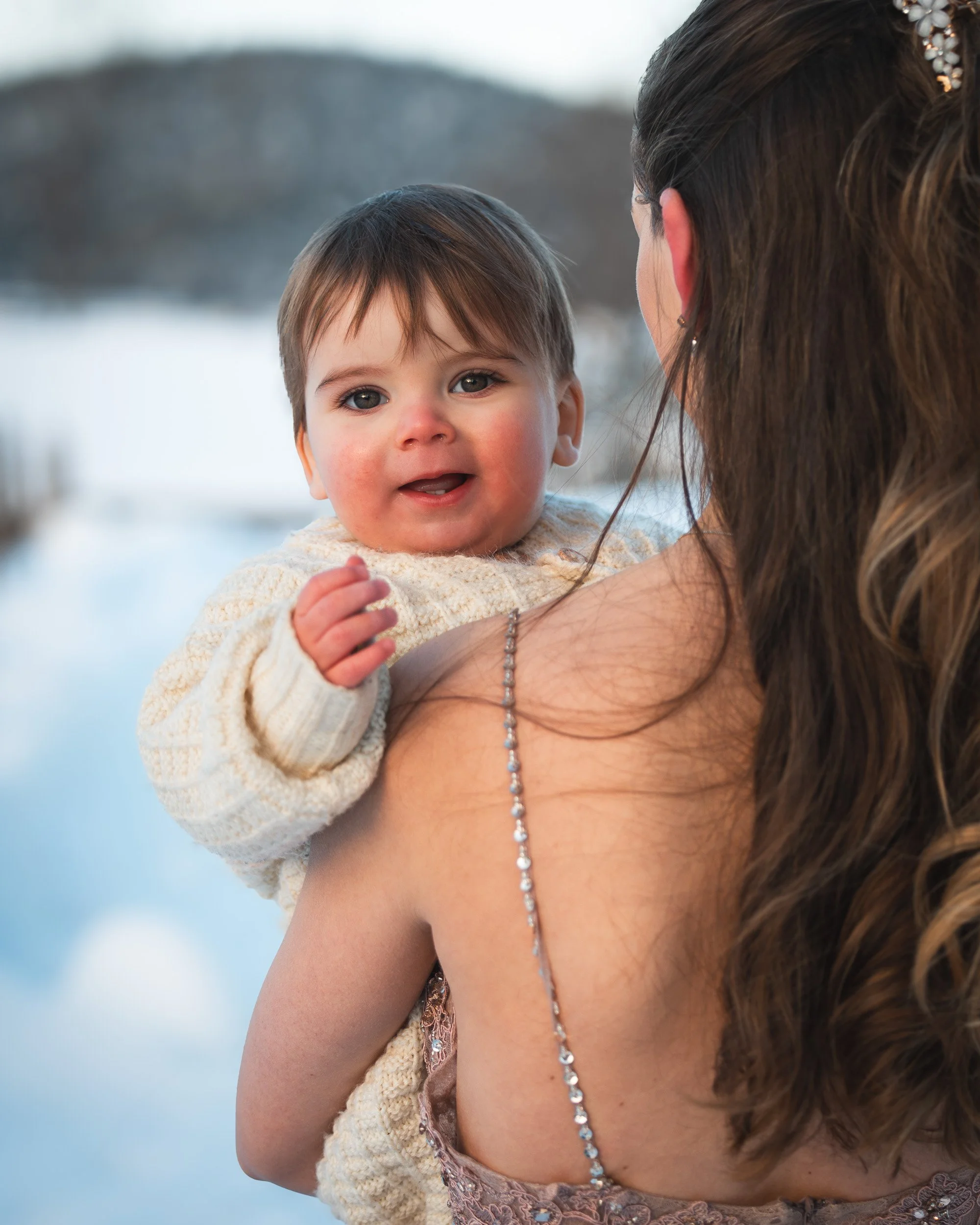 Liten gutt på armen til mamma. Ser i kameraet. Vinterlandskap i bakgrunnen.