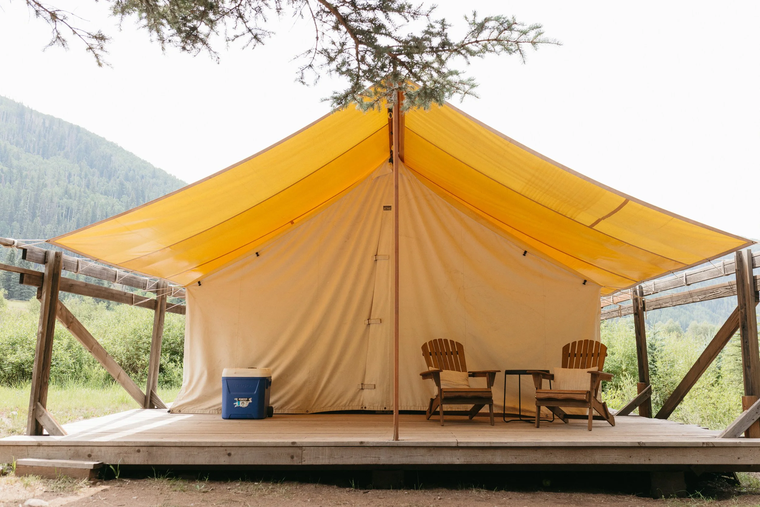 A camping tent on a wooden platform, with two chairs and a small table inside, and a blue cooler outside, set in a mountainous area with green trees and a cloudy sky.
