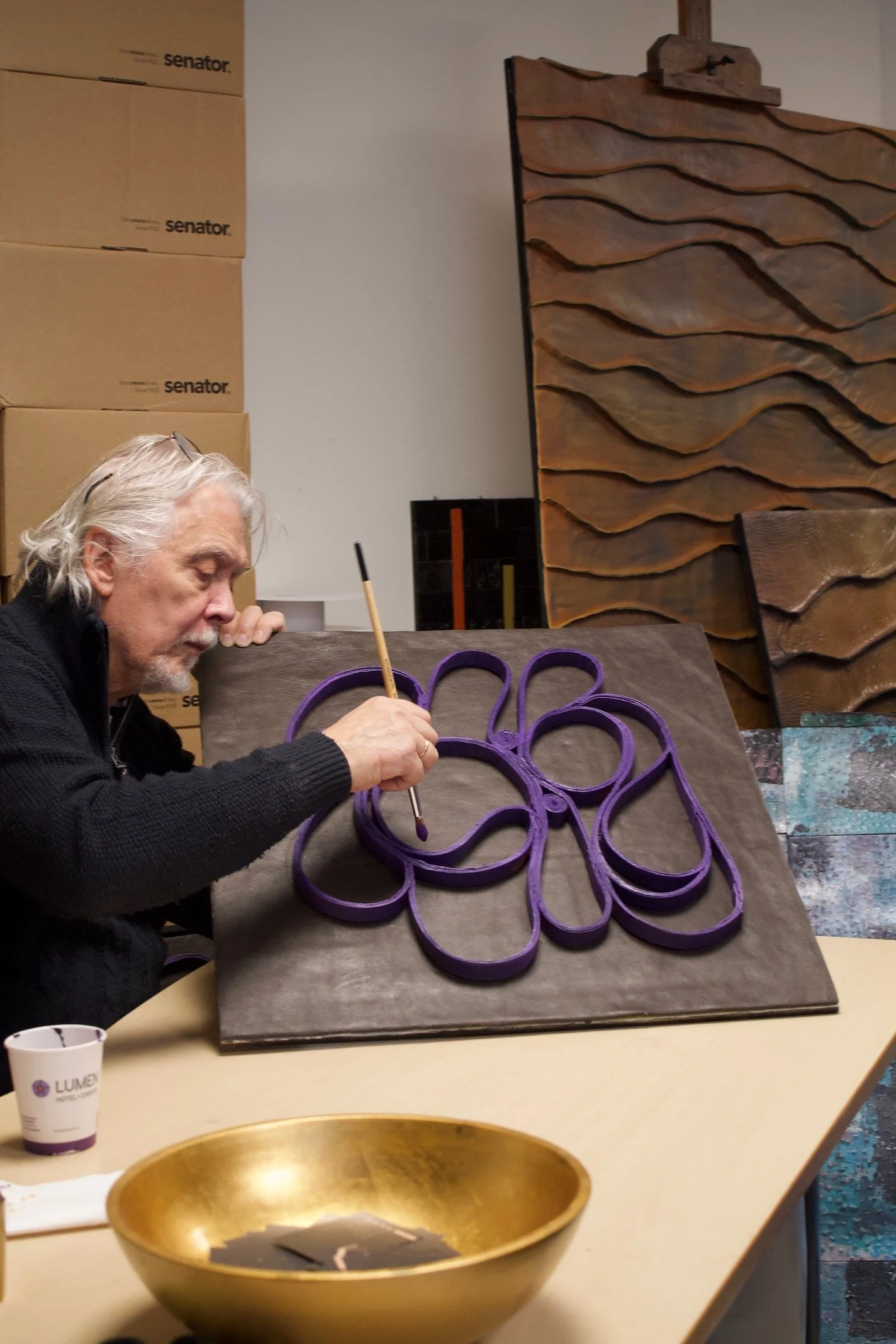 An elderly man with gray hair and a beard is painting a large, abstract artwork on a wooden panel. The artwork features interconnected loops painted purple. The man is using a fine brush, and the scene is set in an artist's studio with various textured wooden panels and cardboard boxes in the background.