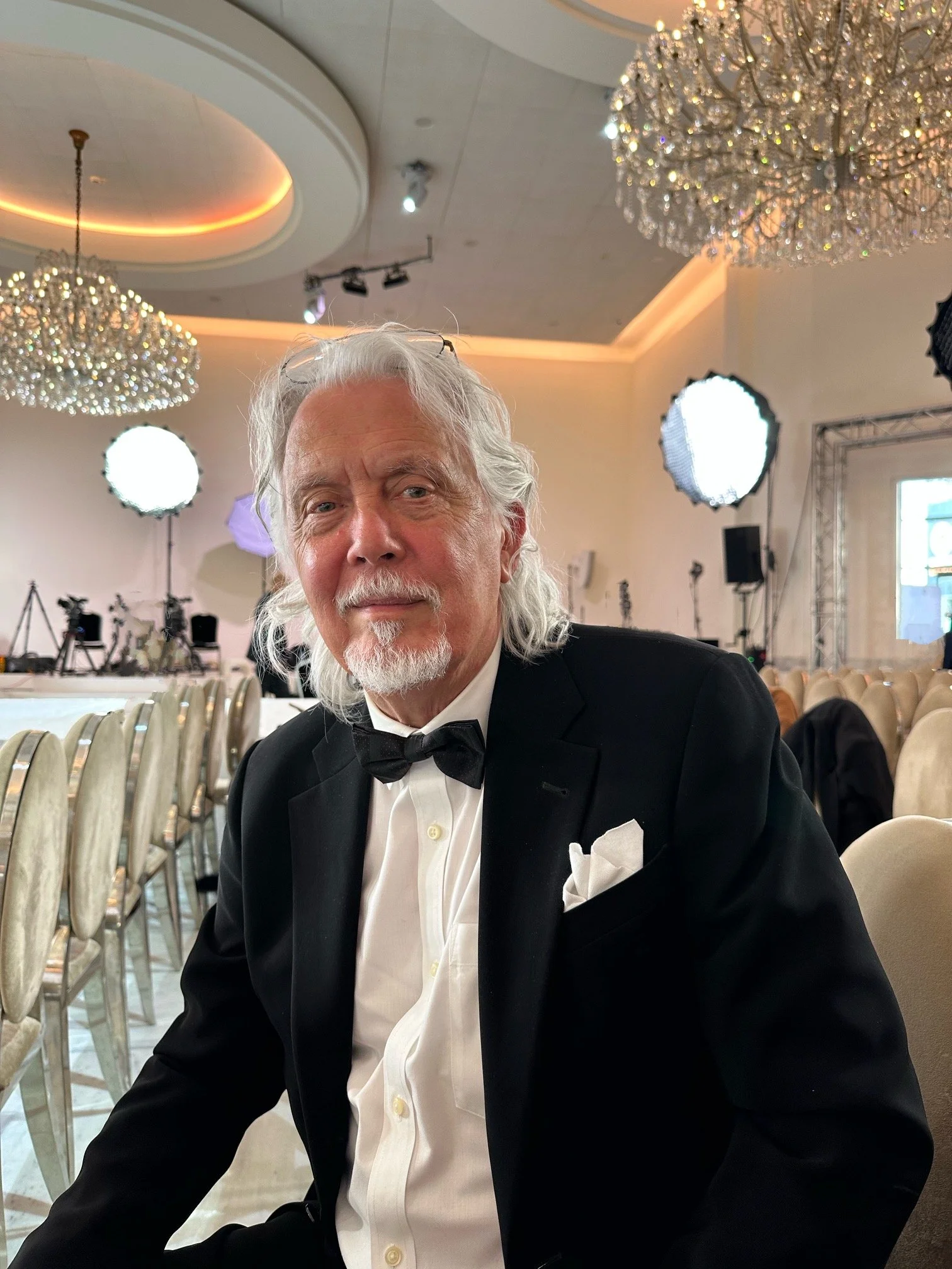 An elderly man with long white hair and a beard, dressed in a black tuxedo with a white shirt, black bow tie, and a white pocket square, sitting at a formal event with chandeliers, stage lights, and rows of chairs behind him.