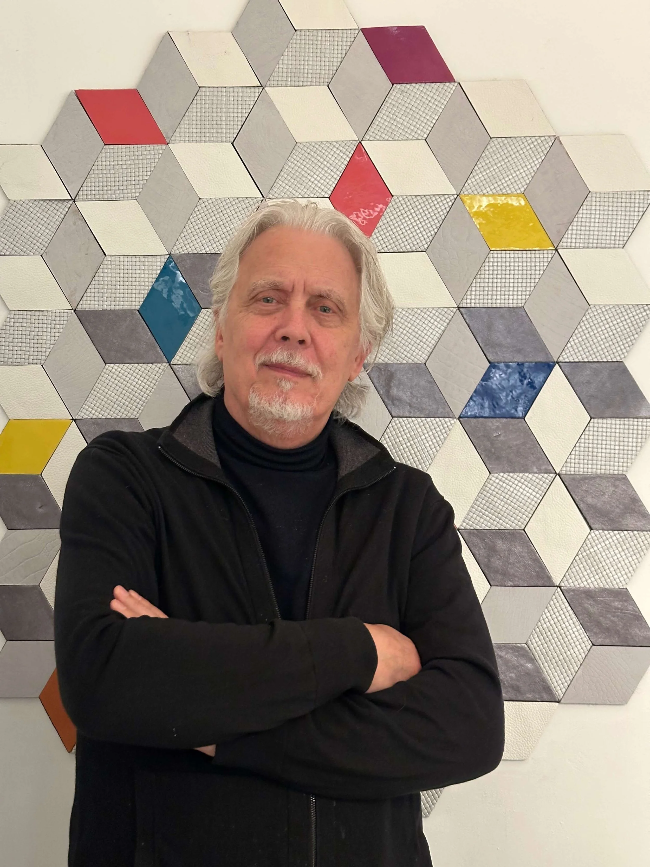A man with gray hair and a beard standing in front of a geometric wall art made of hexagonal tiles, some of which are colored in red, yellow, blue, and orange, with crossed arms and slight smile.