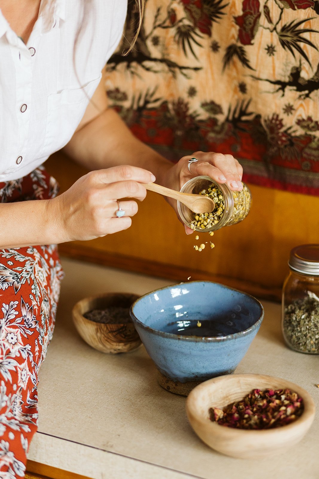 Person adding dried herbs or flowers from a jar into a blue ceramic bowl on a table with other bowls and jars containing herbs or flowers.