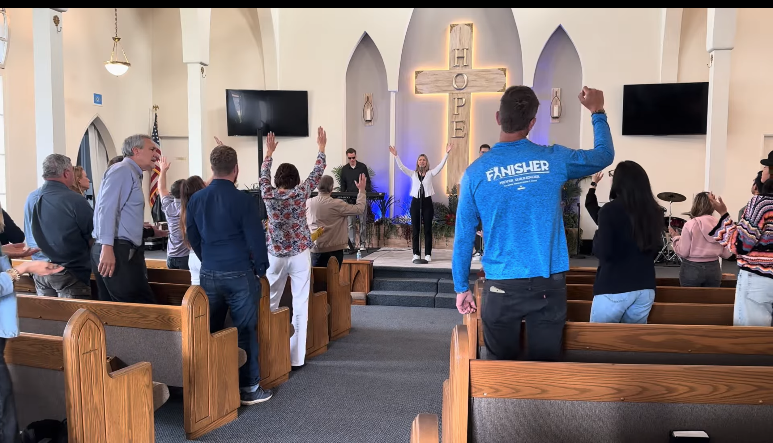 Congregation and choir members worshiping in a church with a large cross on the wall behind the stage, with some members raising their hands, during a religious service or prayer.