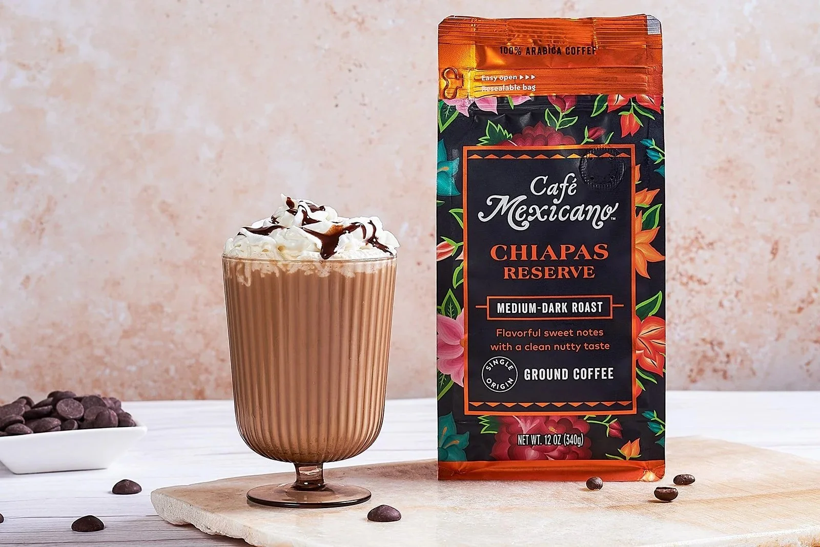 A glass of iced coffee topped with whipped cream and chocolate syrup next to a package of Café Mexicano Chiapas Reserve ground coffee, with a small bowl of coffee beans on a white surface against a beige textured background.