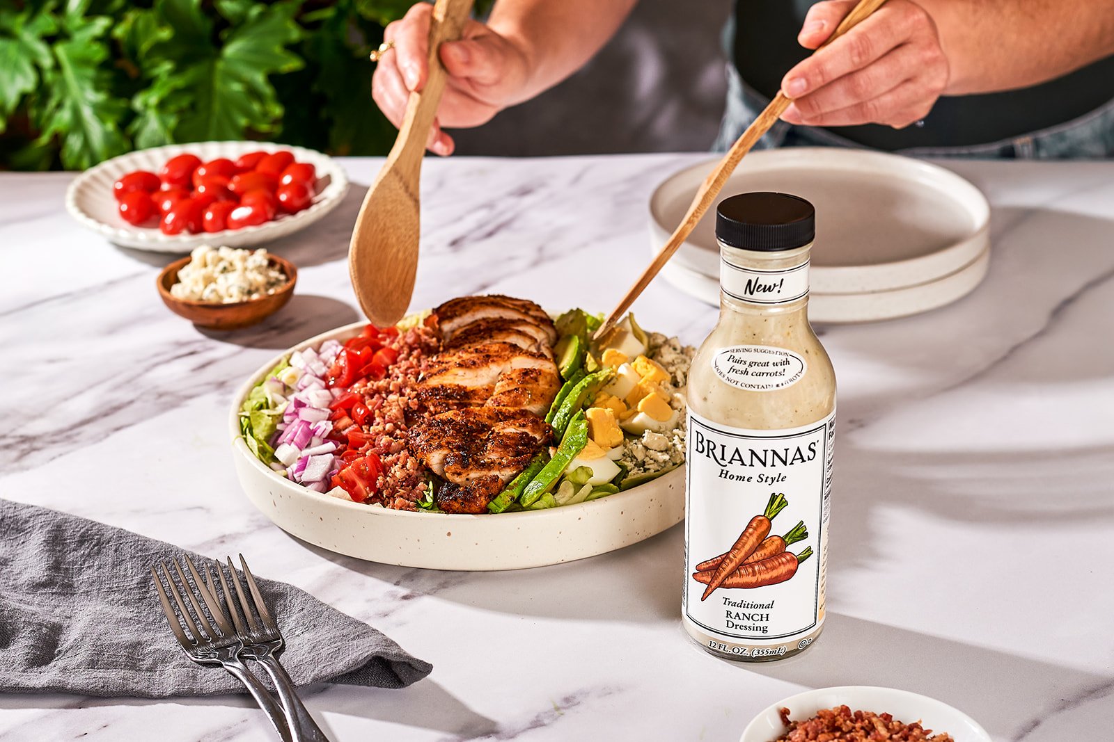 A bowl of salad with sliced grilled chicken, chopped tomatoes, onions, green beans, and crumbled cheese on a white marble table, with a bottle of Brianna's ranch dressing nearby. In the background, there are small bowls of cherry tomatoes and crumbled cheese, and a fork and a napkin on the table.