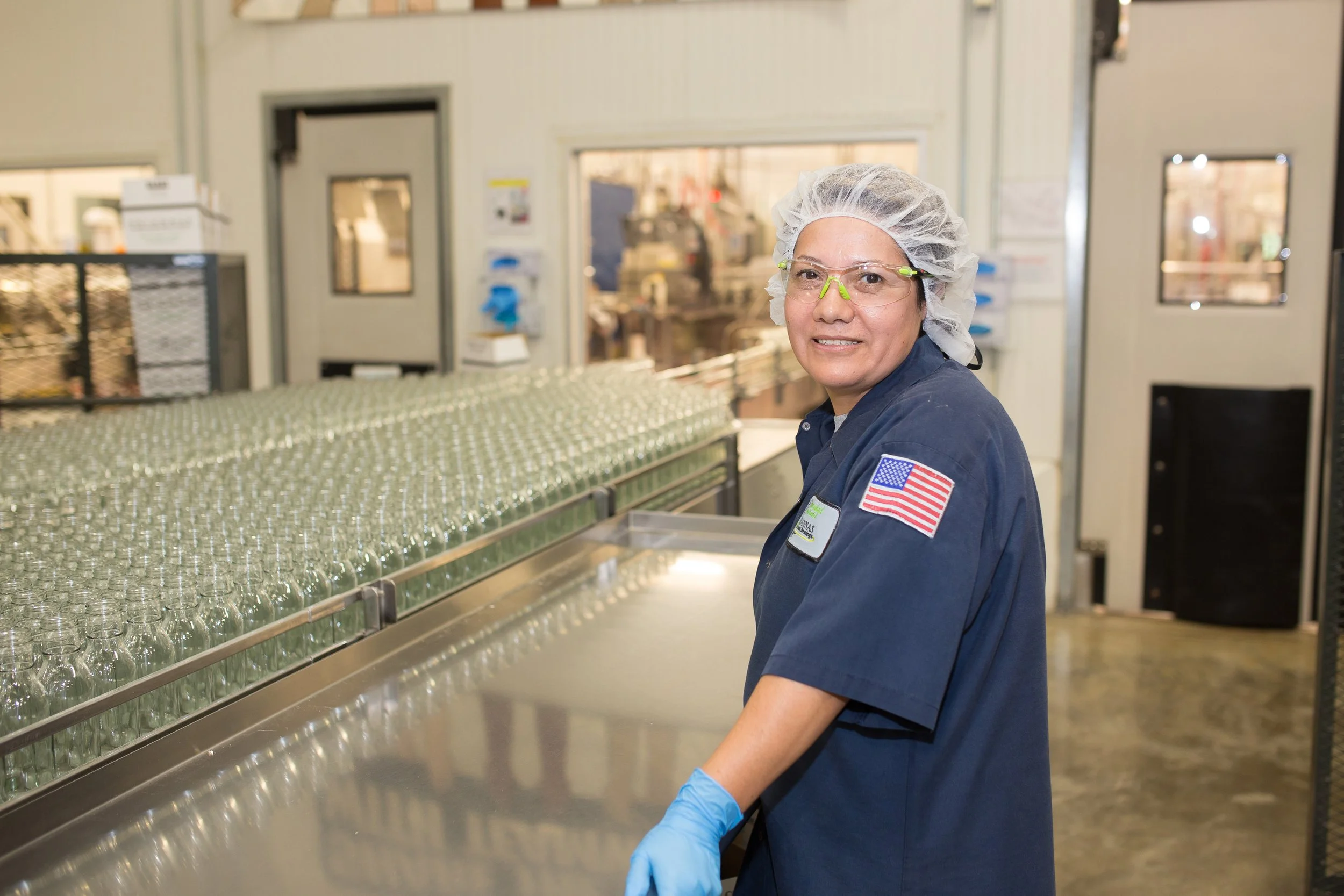 A woman wearing a navy uniform, safety glasses, a hairnet, and blue gloves stands in a glass manufacturing facility with glass bottles on a production line.