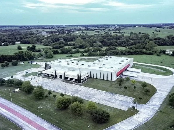 Large industrial or commercial building with a parking lot, surrounded by green trees and open fields under a partly cloudy sky.