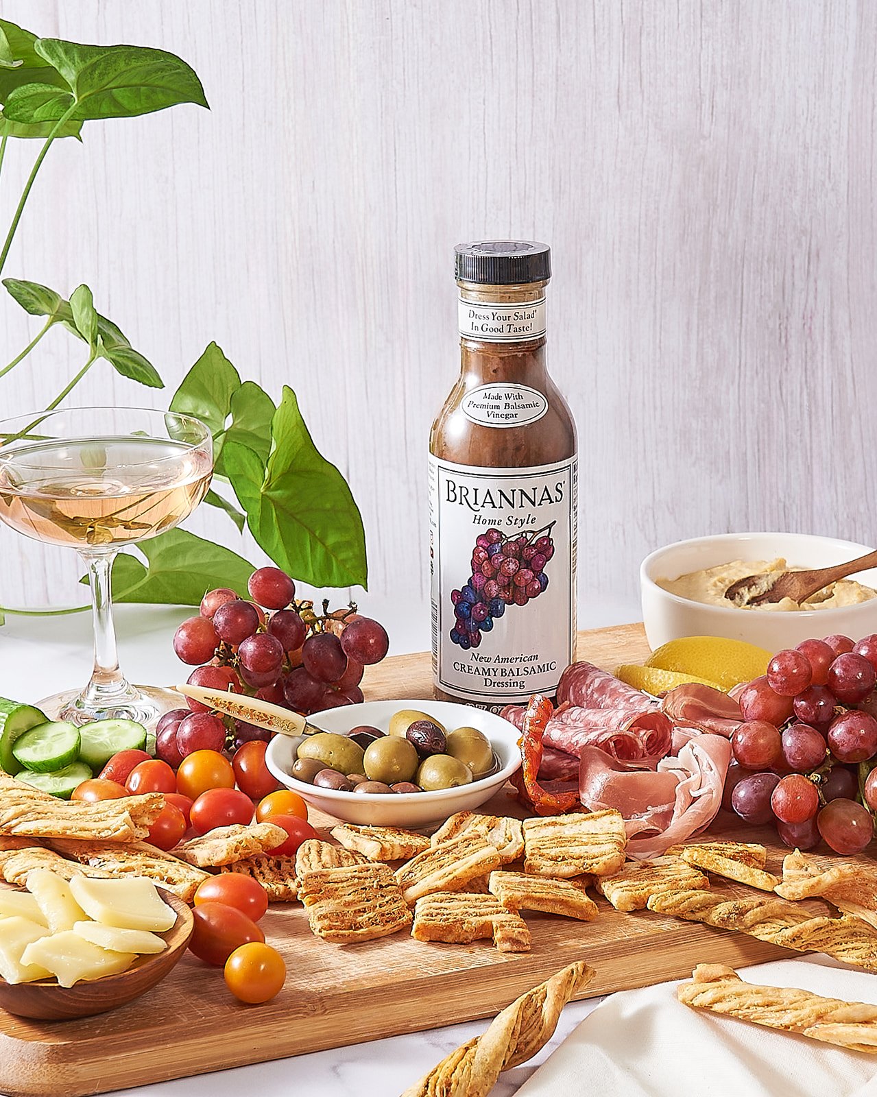 A charcuterie board with cheeses, grapes, crackers, cherry tomatoes, olives, and a bottle of Brianna's creamy balsamic dressing, alongside a glass of rosé wine and a bowl of salad dressing.