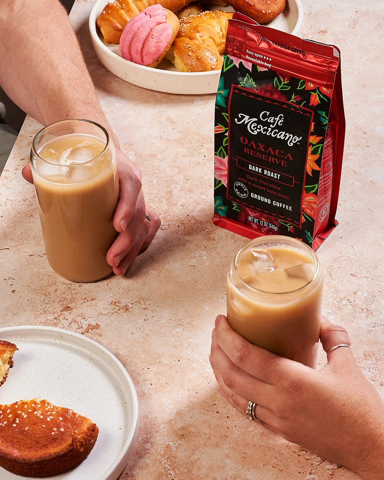 Two people holding glasses of iced coffee, a bag of Café Mexicano Oaxaca Reserve dark roast ground coffee, a plate of partially eaten baked goods, and a bowl of assorted cookies on a table.