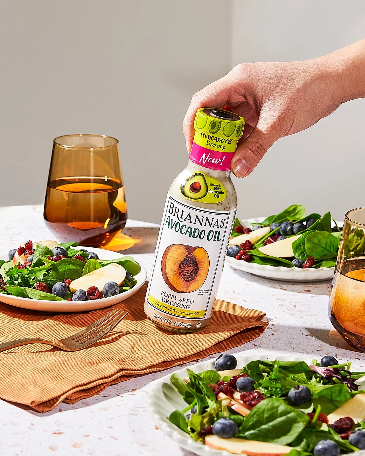 A hand holding a bottle of Brianna's Avocado Oil Poppy Seed Dressing over a table with salads, glasses of brown drinks, and an orange napkin.