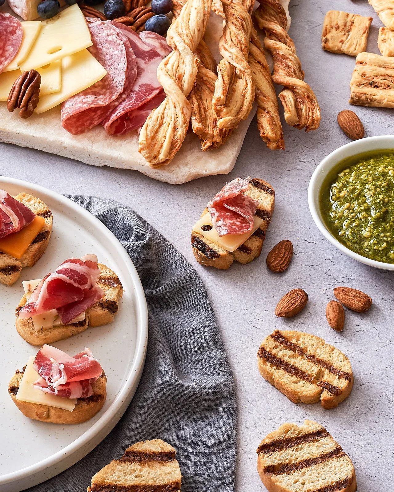 A charcuterie board with sliced cheeses, cured meats, bread, nuts, and a green sauce, with crostini topped with cheese and meats, all on a light gray surface and a gray napkin.