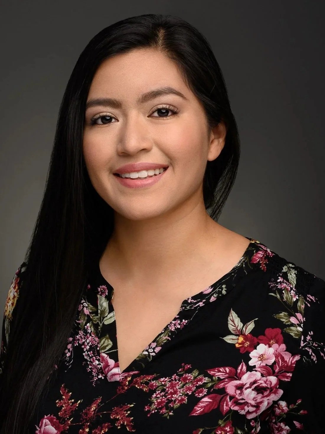 A professional headshot of Paralegal, Isabel Campos-Axilote, a woman with long dark hair, smiling, wearing a black floral blouse with pink and red flowers, against a dark gray background.