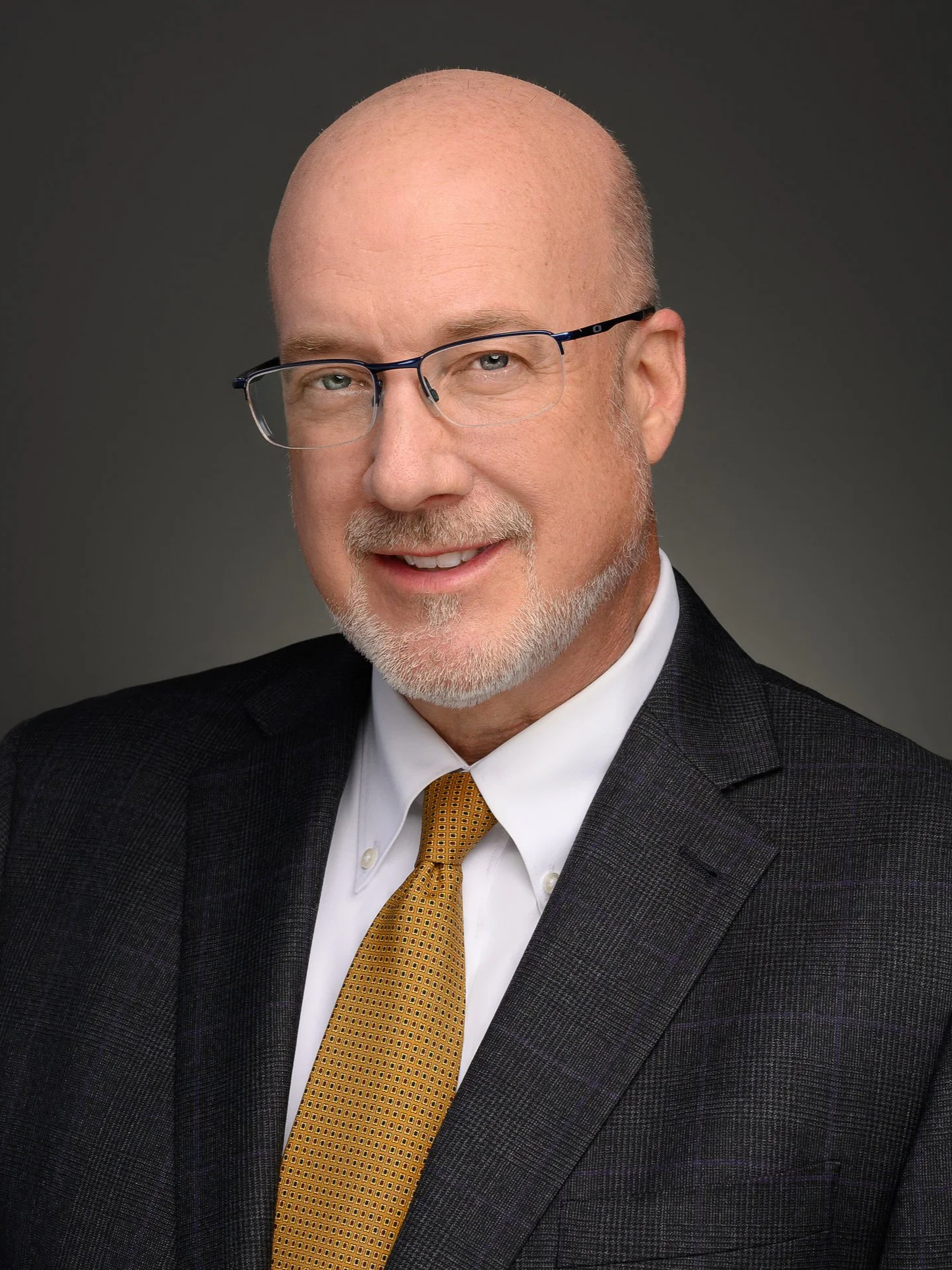 A professional headshot of Senior Partner, Robert C. Elfont, a middle-aged man with glasses, a beard, wearing a dark suit, white shirt, and a yellow tie, against a plain dark background.