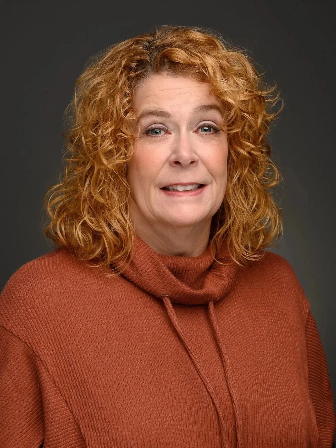 A professional headshot of Senior Legal Assistant / Client Navigator, Barbara M. Guendelsberger, a woman with curly red hair wearing an orange turtleneck sweater, smiling slightly, against a dark gray background.