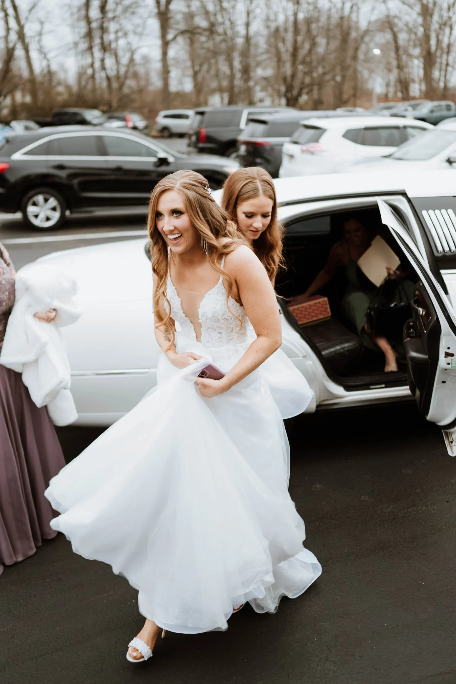 A bride in a white wedding dress stepping out of a limousine, smiling, with bridesmaids around her, in a parking lot.