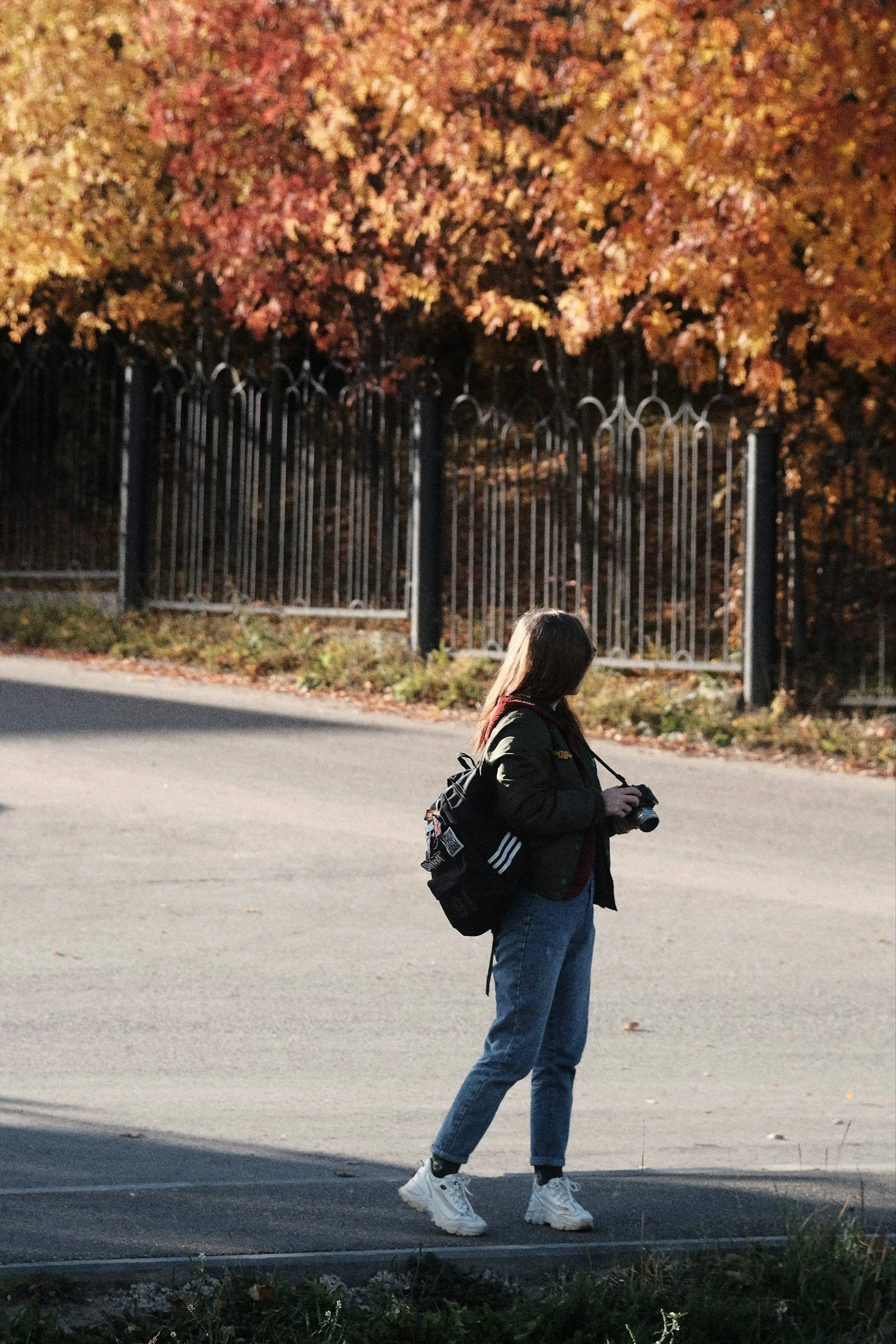 A young woman standing on the sidewalk holding a camera, with autumn colorful trees and iron fence in the background.