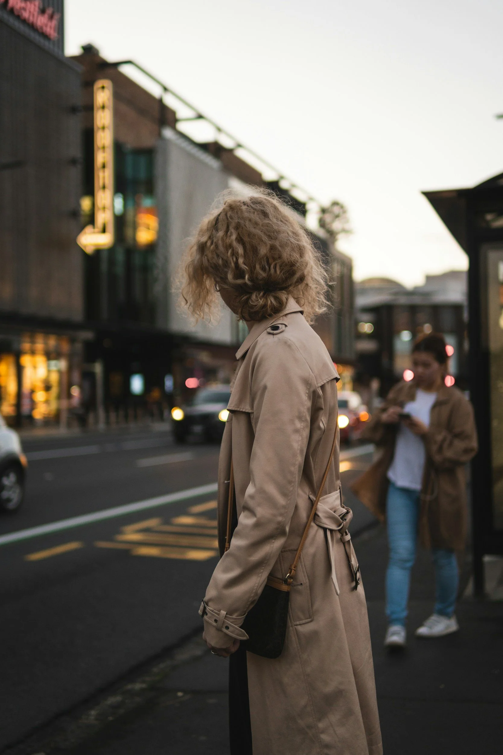 A woman with curly blonde hair wearing a tan trench coat standing on a city sidewalk at dusk, with a woman in the background looking at her phone, and cars on the street.