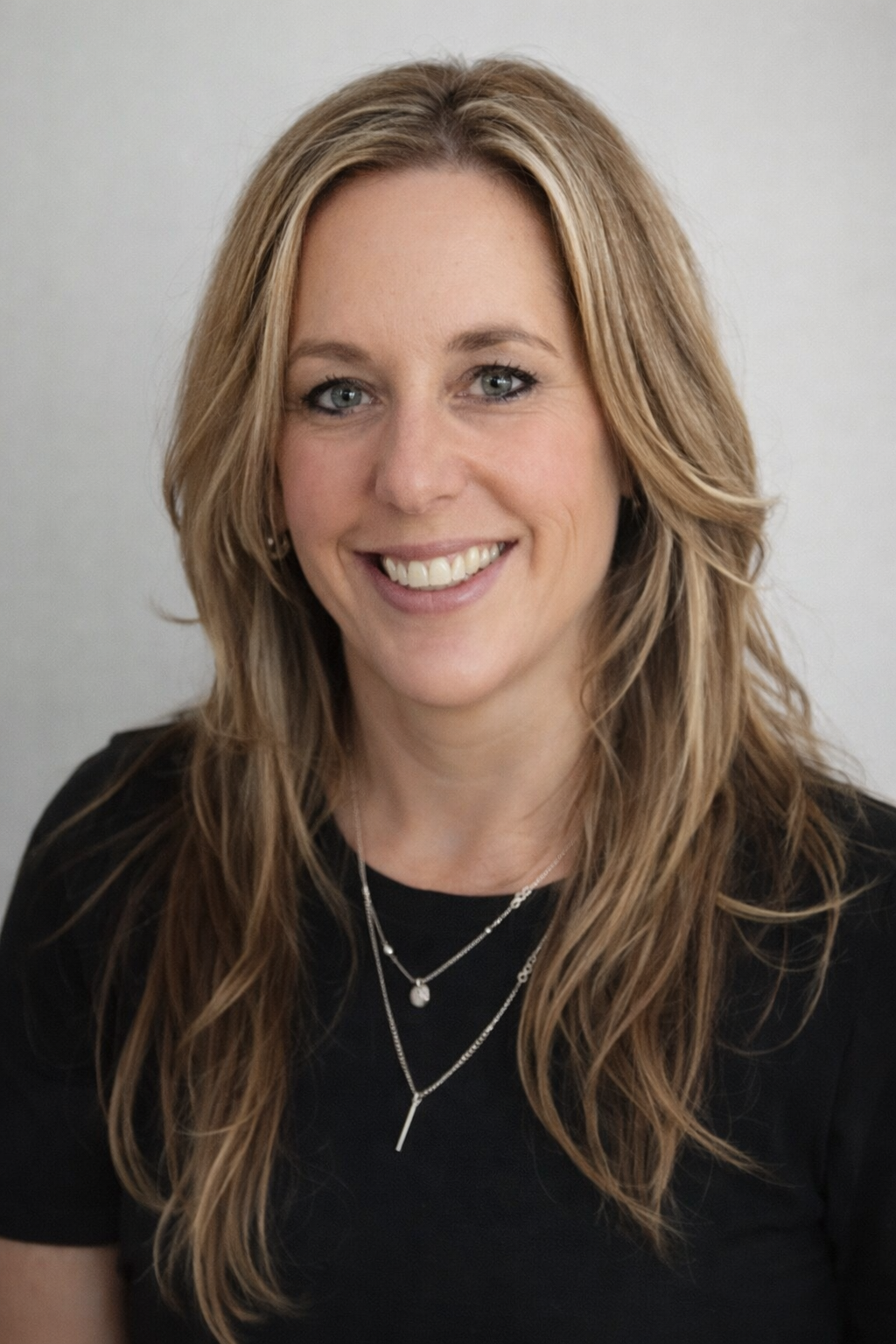 A woman with long, wavy blonde hair and blue eyes, smiling while wearing a black top and layered silver necklaces, against a plain light-colored background.
