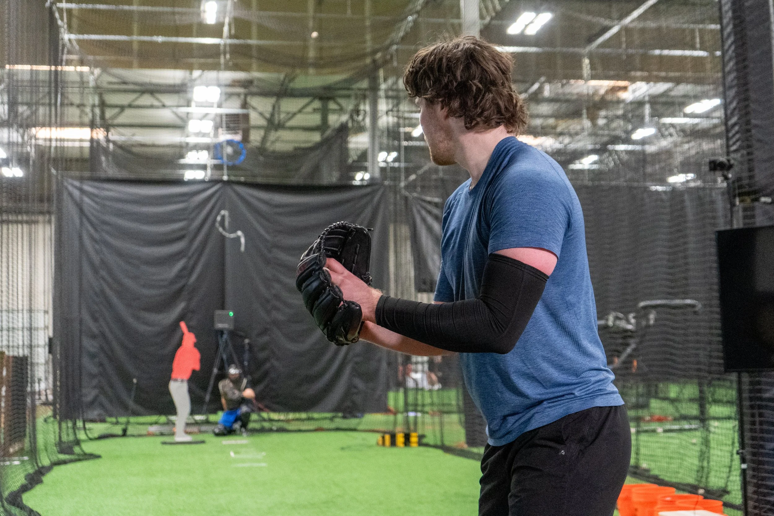 A young man in a blue t-shirt and black pants wearing a black baseball glove and a black arm sleeve, standing in an indoor baseball or softball training facility.