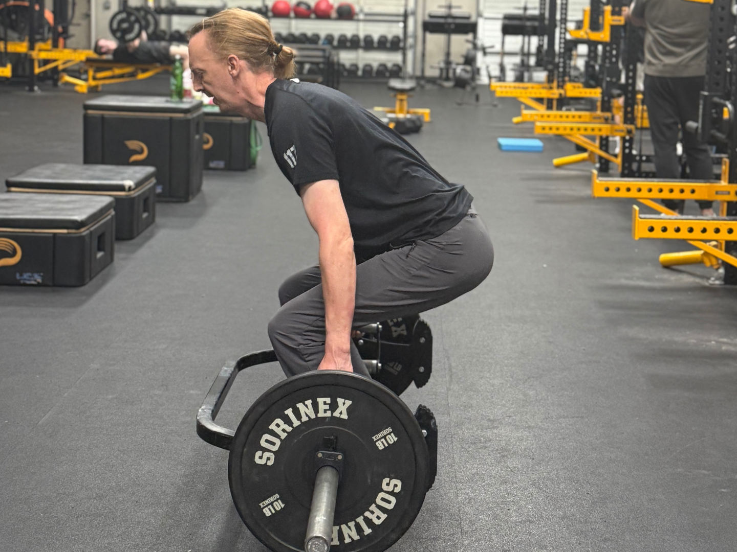 A man with long hair tied back, wearing a black t-shirt and gray pants, is lifting a barbell with weight plates in a gym.