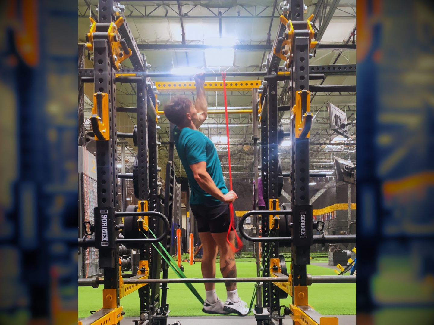 A man in a teal shirt and black shorts performing a pull-up exercise in a gym with a black and yellow power rack, fitness equipment, and bright lighting.