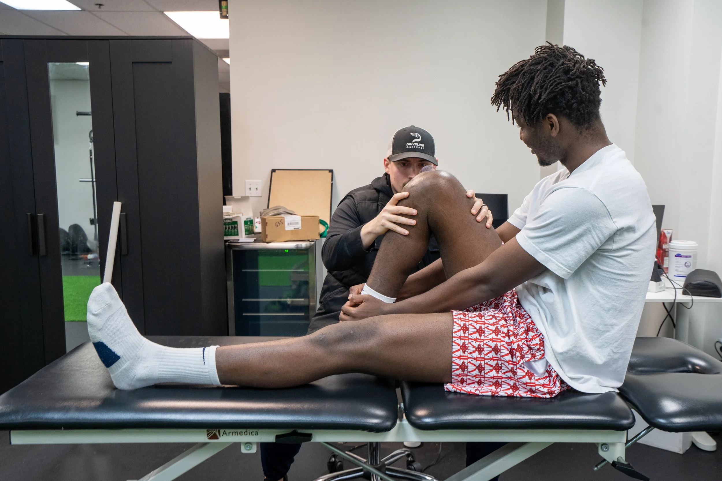 A physical therapist or trainer assists a young man with a leg stretch or exercise, sitting on an examination table in a clinical or physical therapy setting.