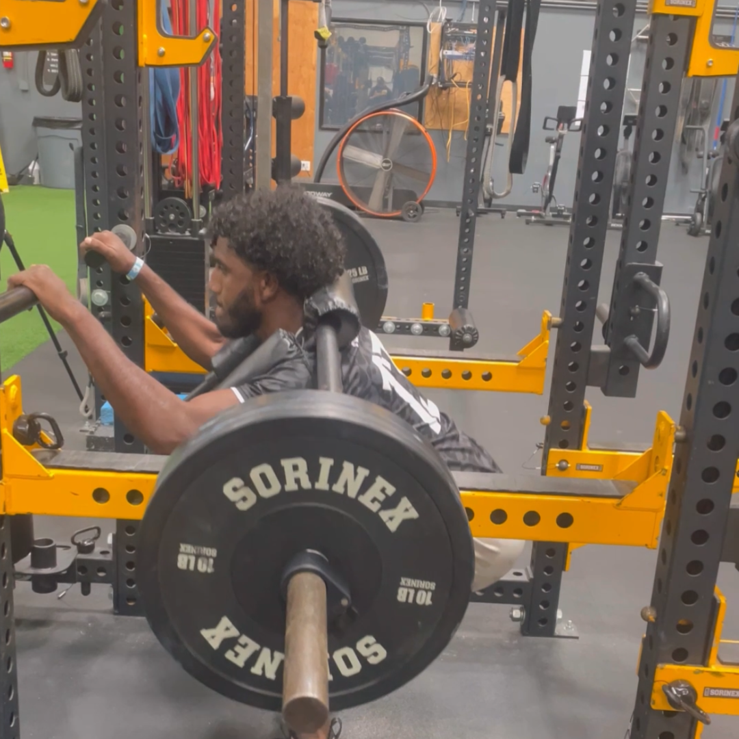 Man performing a barbell bench press in a gym, surrounded by workout equipment and weights.