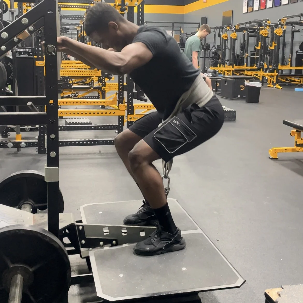 A man with a chain around his waist performing a squat exercise in a gym, using a squat rack, with a person in the background working out and gym equipment visible.