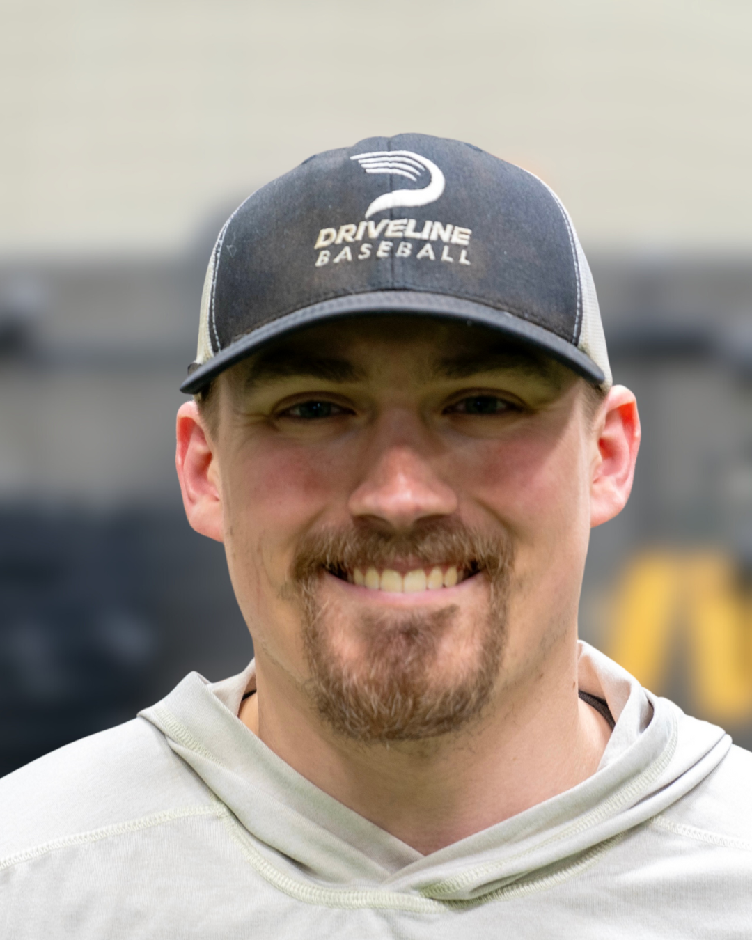 A man wearing a black baseball cap with 'Driveline Baseball' logo and a light-colored hoodie, smiling indoors.