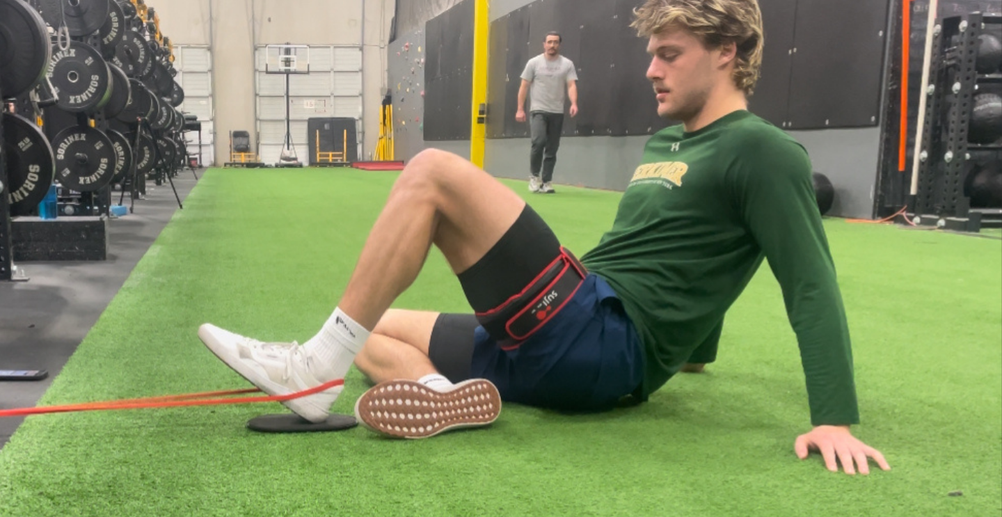 A young man exercising on a gym floor with a resistance band and a trainer walking in the background.