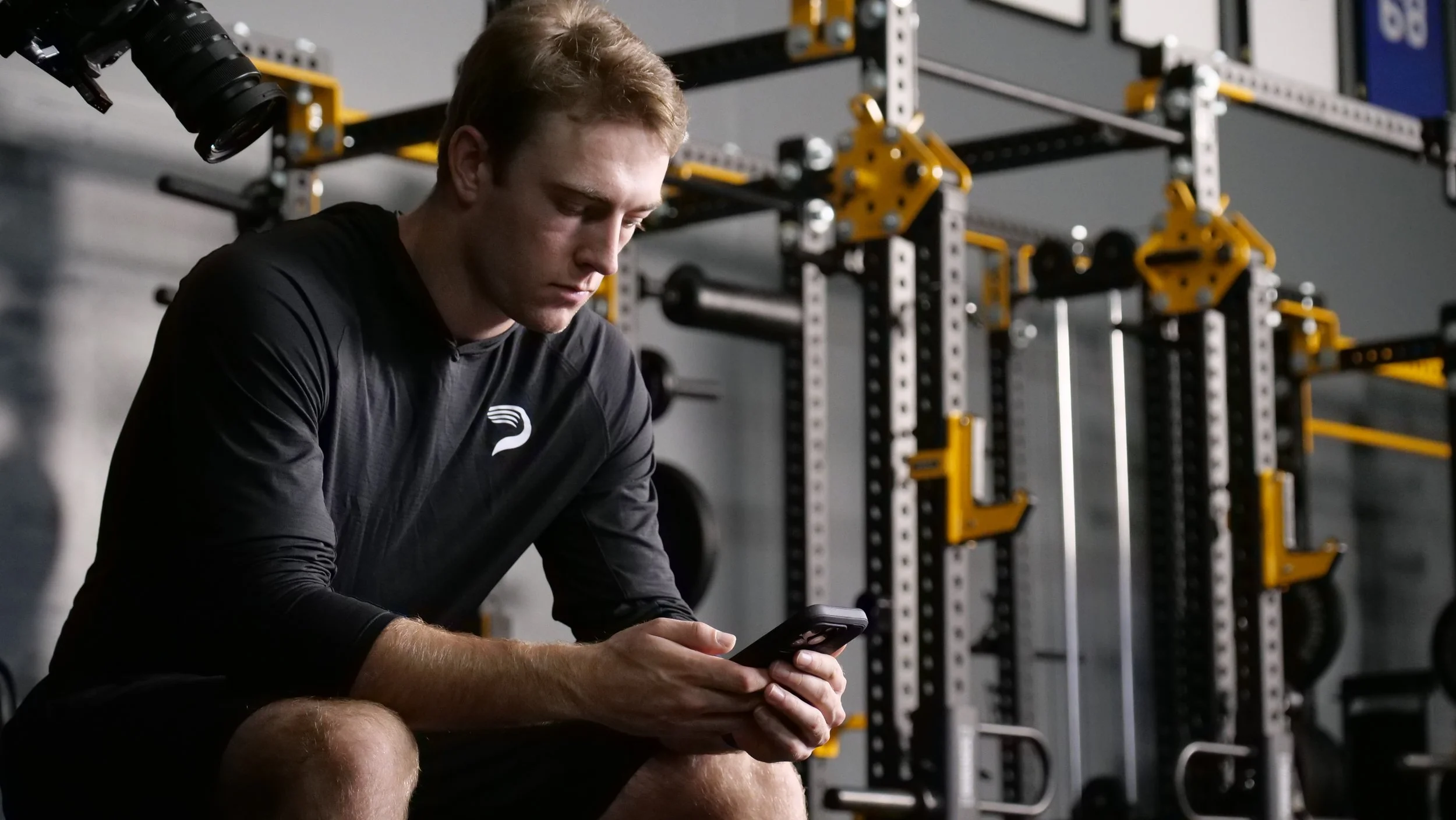 A man in athletic clothing sitting in a gym, looking at his phone with a squat rack and gym equipment in the background.