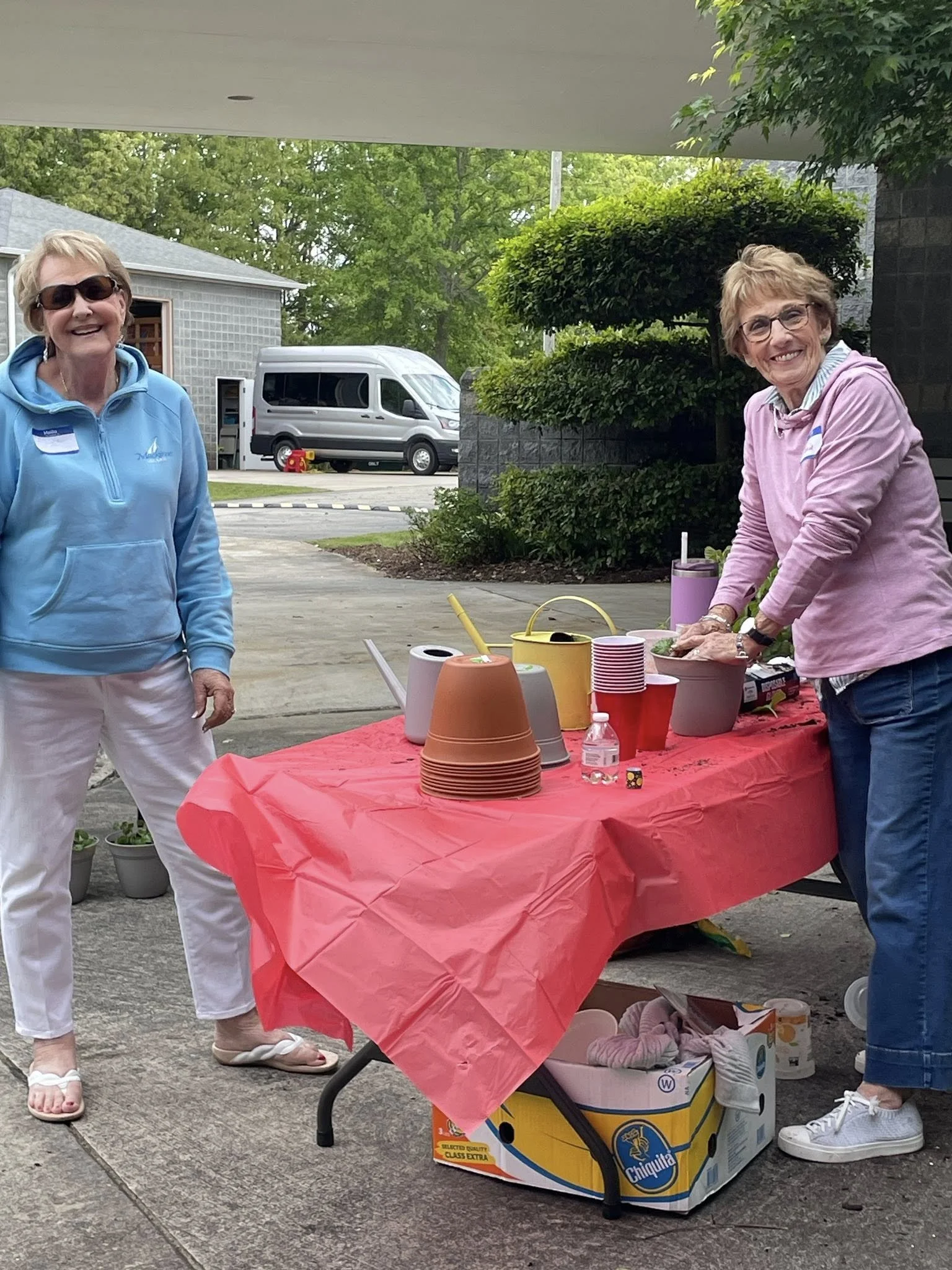 Two women standing at a table covered with a red tablecloth, outdoor setting. The table has disposable cups, a water bottle, and buckets, with a box of Chiquita bananas underneath.