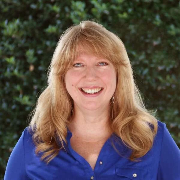 A woman with blonde hair, blue eyes, and a bright smile wearing a blue shirt outdoors with greenery in the background.