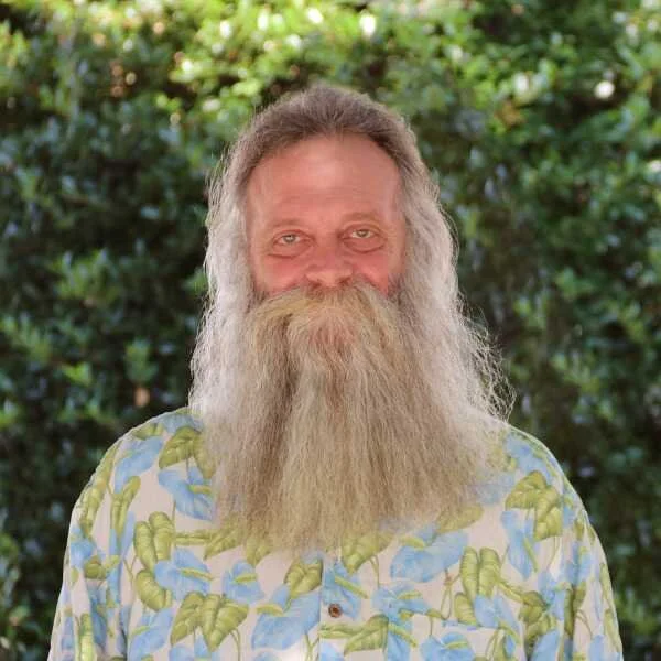 A bearded man smiling outdoors with a background of green foliage, wearing a colorful Hawaiian shirt with a leafy pattern.