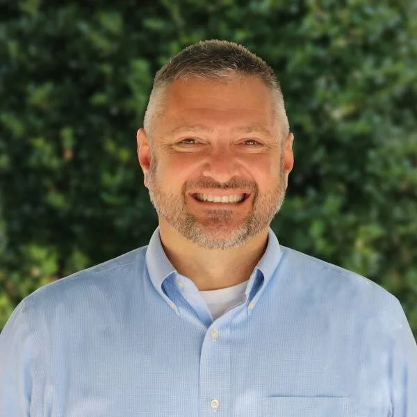 A man with short gray hair and a beard smiling outdoors, wearing a light blue button-up shirt against a background of green trees.
