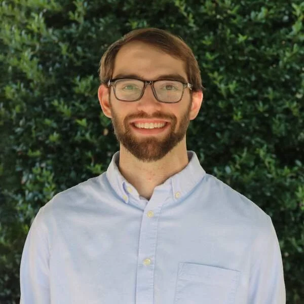 A young man with glasses and a beard, smiling outdoors in front of greenery, wearing a light blue button-up shirt.