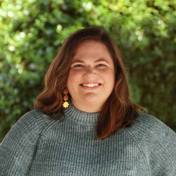 A woman with shoulder-length brown hair, wearing a gray sweater and colorful flower-shaped earrings, smiling outdoors with greenery in the background.