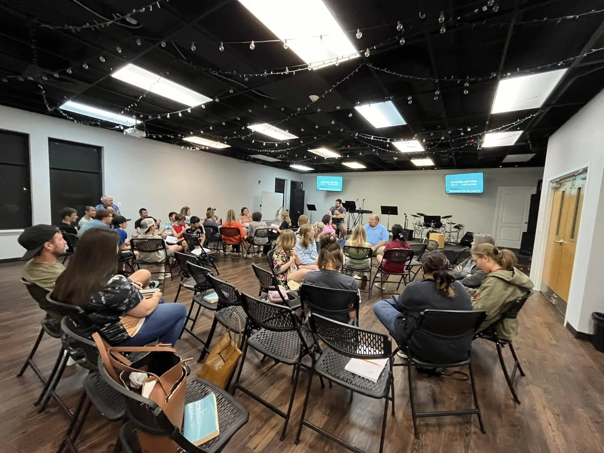 A group of people seated in chairs facing a stage with a person speaking into a microphone. The stage has musical instruments and two screens displaying blue background visuals. The room has black ceiling and string lights, with wood flooring.