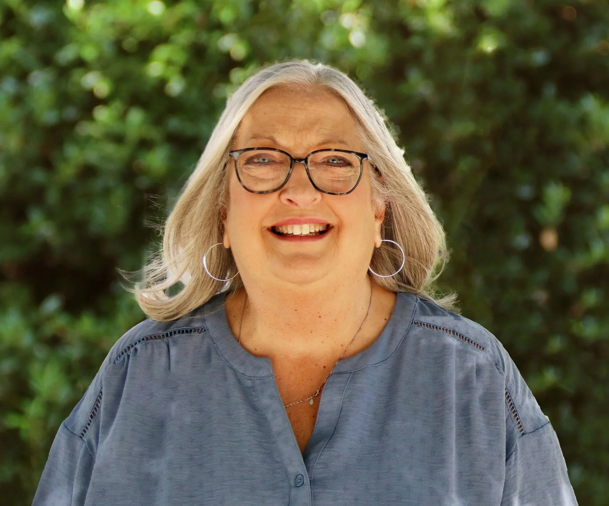 A smiling older woman with gray hair, glasses, and hoop earrings standing outdoors with green foliage in the background.