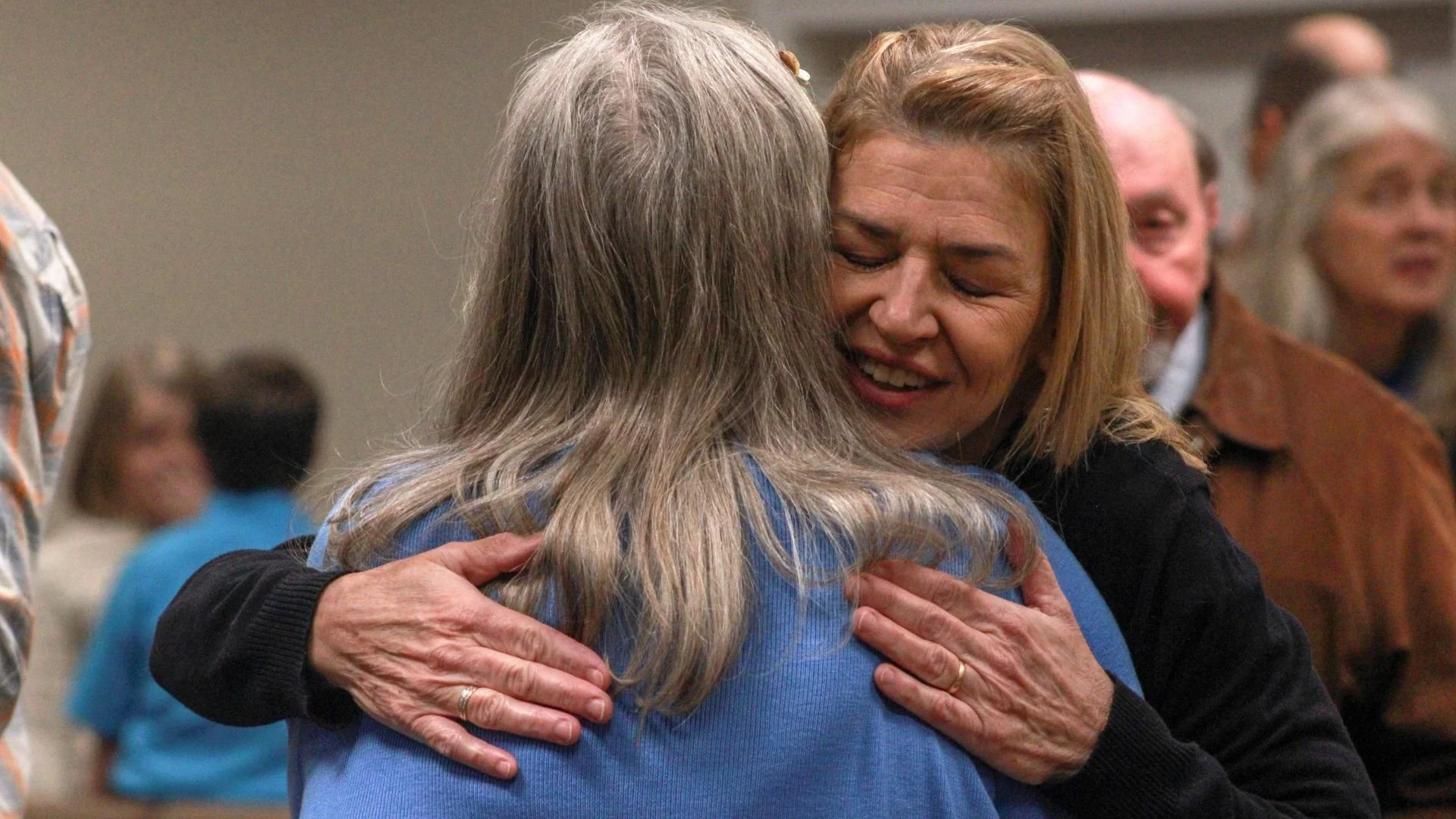 Two women hugging each other warmly in a crowded room, with one woman smiling with her eyes closed.