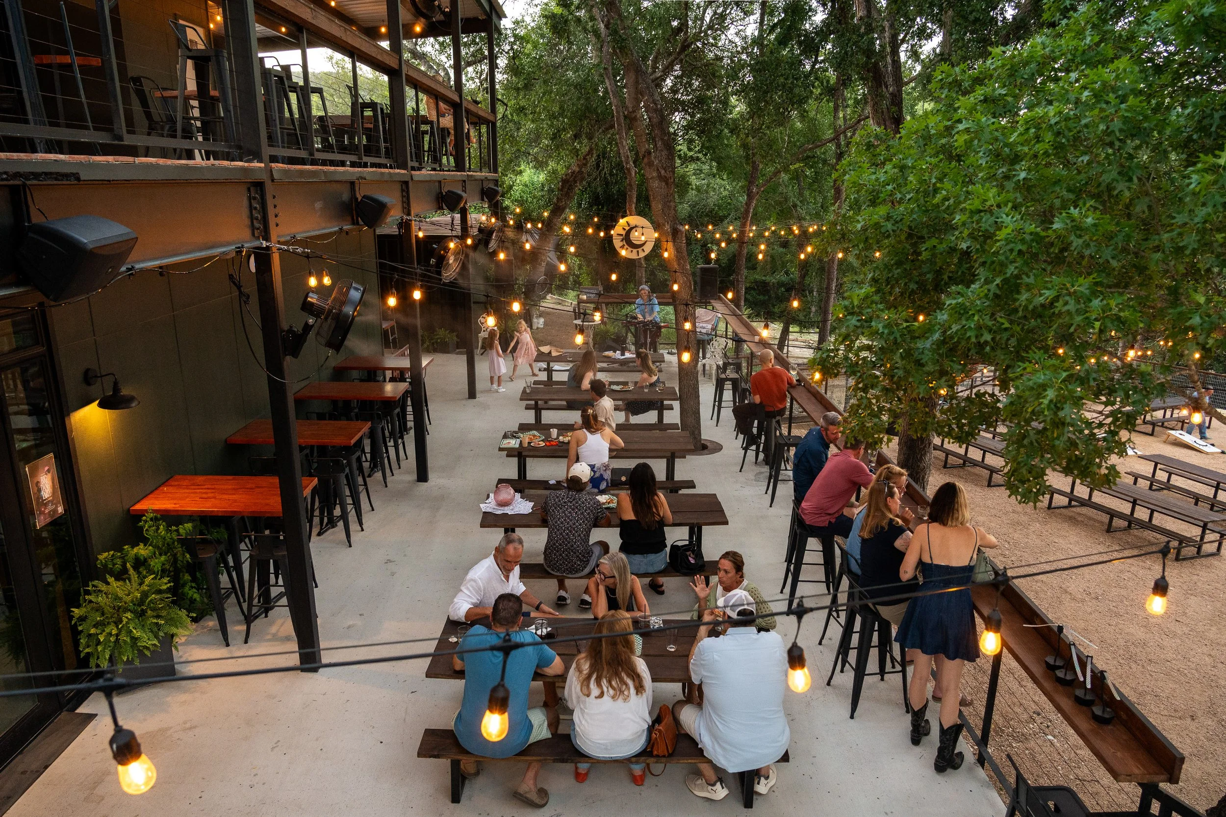 Multiple groups of people sitting at wooden tables and standing in an outdoor patio area decorated with string lights and trees, with a building featuring a balcony and some children dancing in the background.
